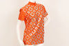 Orange, short-sleeve, mock-neck shirt with a silver, graphic print with mirrored embellishments. 