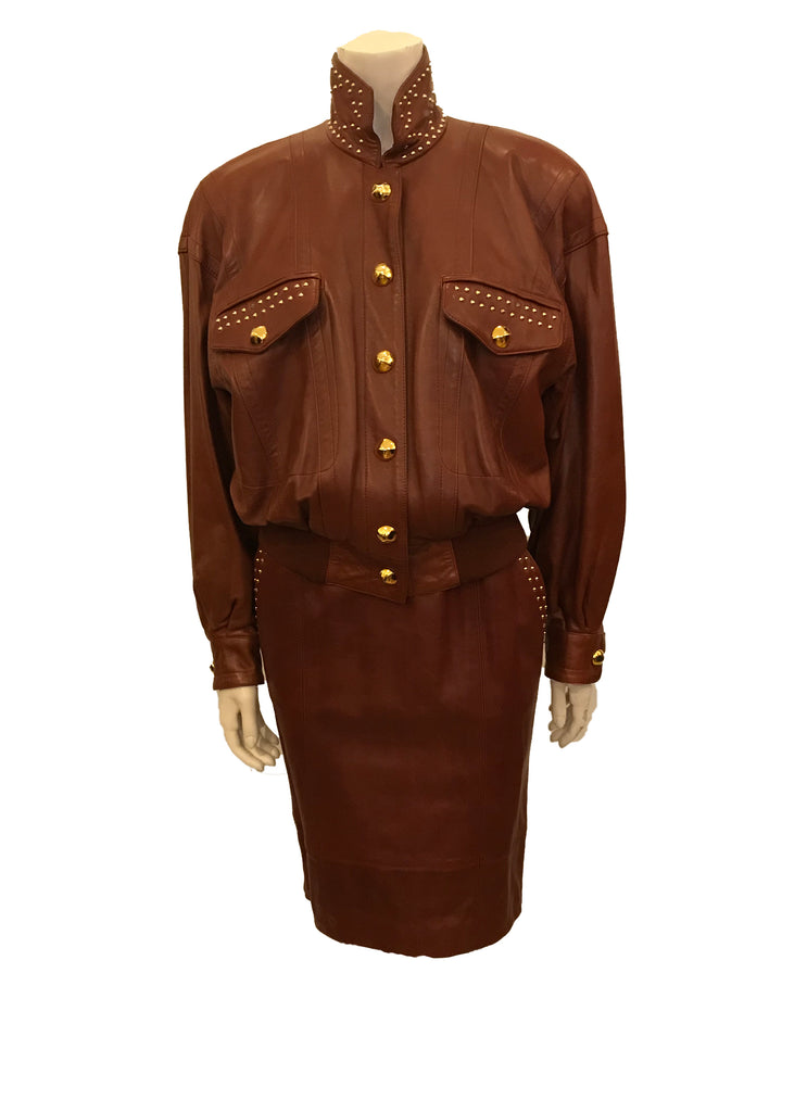 Front view on mannequin a two piece skirt and jacket in burgundy leather Jacket has gold studs on collar and front pockets. Skirt is straight and knee length. Jacket closes with six gold buttons