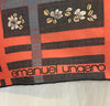Closeup of designer's name "emanuel ungaro" in lower corner of Emanuel Ungaro scarf with a black background and oversize windowpane plaid motif in coral and grey-blue. In the squares of the plaid, there are small clusters of blue and white flowers.