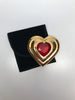 Front view of Yves Saint Laurent gold Heart shaped makeup compact with large red jewel in center sitting next to small black velvet bag included with original packaging. 