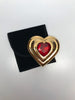Front view of Yves Saint Laurent gold Heart shaped makeup compact with large red jewel in center sitting next to small black velvet bag included with original packaging.